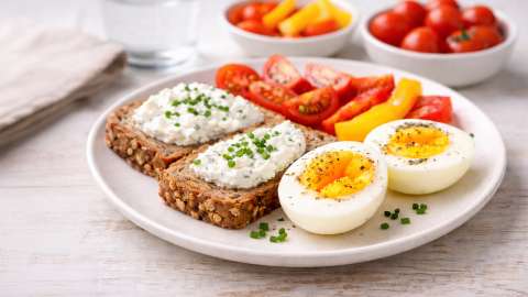 Weiches Ei mit flüssigem Eigelb als Proteinquelle, serviert mit Dinkelvollkornbrot, Frischkäseaufstrich und frischem Gemüse – ausgewogenes Frühstück zur Unterstützung eines aktiven Stoffwechsels.