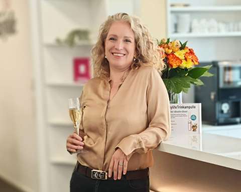 Marie-Stephanie Kolb lächelt in einer goldfarbenen Bluse und hält ein Sektglas in der Hand.