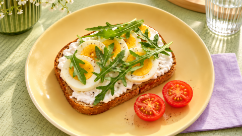 Roggenbrot mit Topfen, gekochten Eierscheiben und Rucola, daneben Tomaten auf einem gelben Teller.